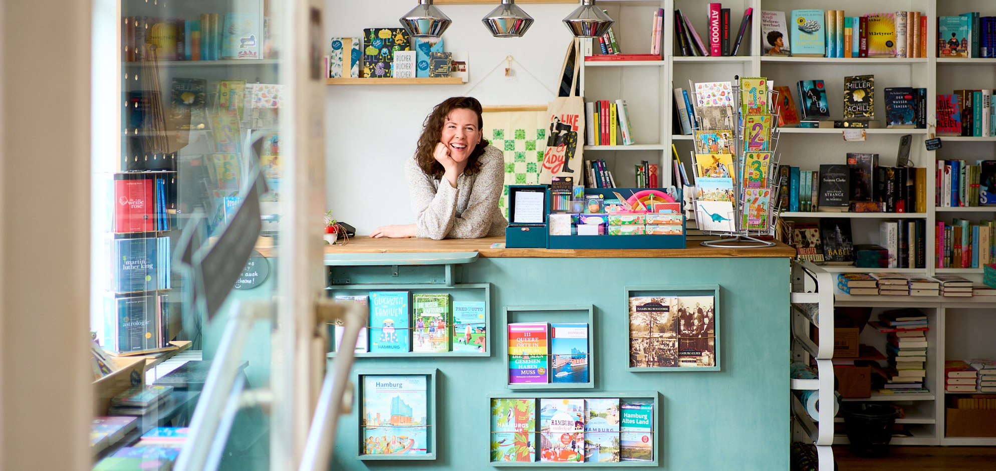 Innenansicht einer einladenden Buchhandlung mit der Besitzerin am türkisfarbenen Verkaufstresen, darauf Bücher und Postkarten. Regale voller Bücher und Pflanzen schmücken den Raum, der helle, moderne Einrichtung und dekorative Beleuchtung bietet. Ein Einblick in die Vielfalt und Kreativität der Buchhandlung.