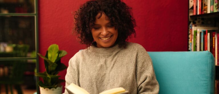 Buchhändlerin Christina Njehu vom Literatourcafé sitzt auf einem türkisfarbenen Sessel in einem gemütlichen Raum mit roten Wänden und hält ein geöffnetes Buch. Neben ihr steht ein kleiner Tisch mit einer Zimmerpflanze darauf. Im Hintergrund sind Bücherregale zu sehen.