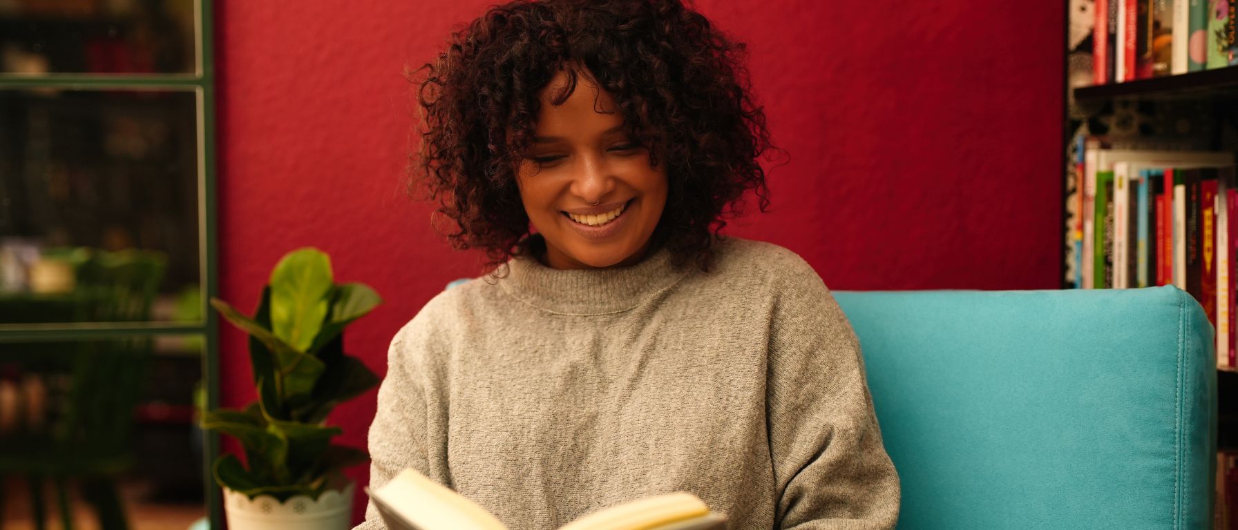 Buchhändlerin Christina Njehu vom Literatourcafé sitzt auf einem türkisfarbenen Sessel in einem gemütlichen Raum mit roten Wänden und hält ein geöffnetes Buch. Neben ihr steht ein kleiner Tisch mit einer Zimmerpflanze darauf. Im Hintergrund sind Bücherregale zu sehen.